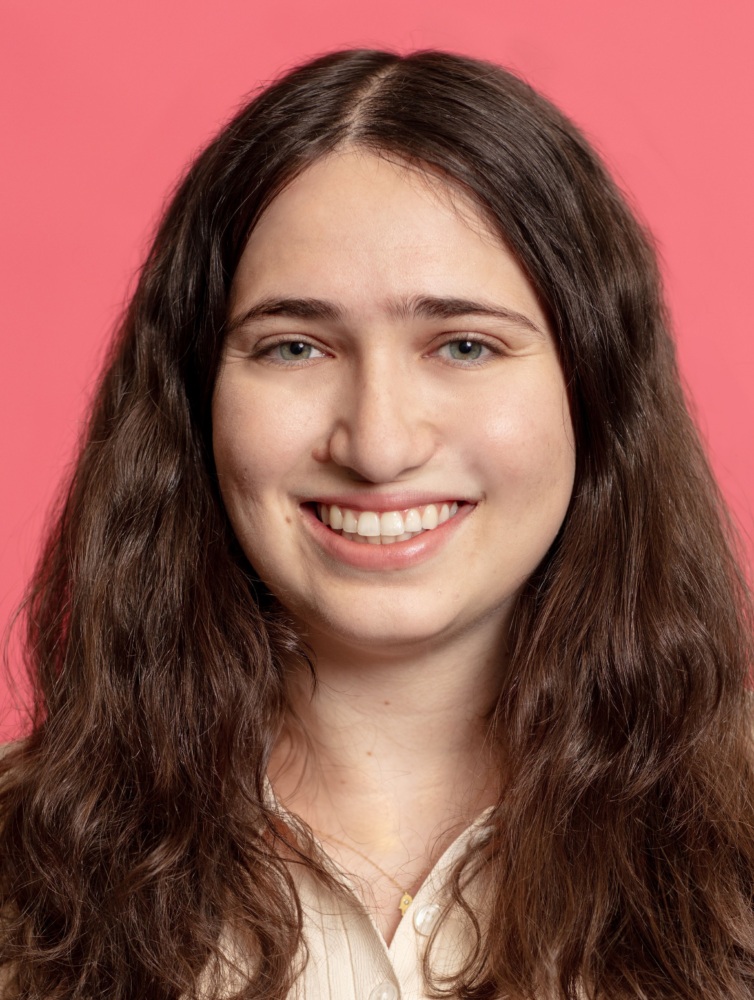 A person with long brown hair smiles in front of a pink background. They are wearing a white shirt.